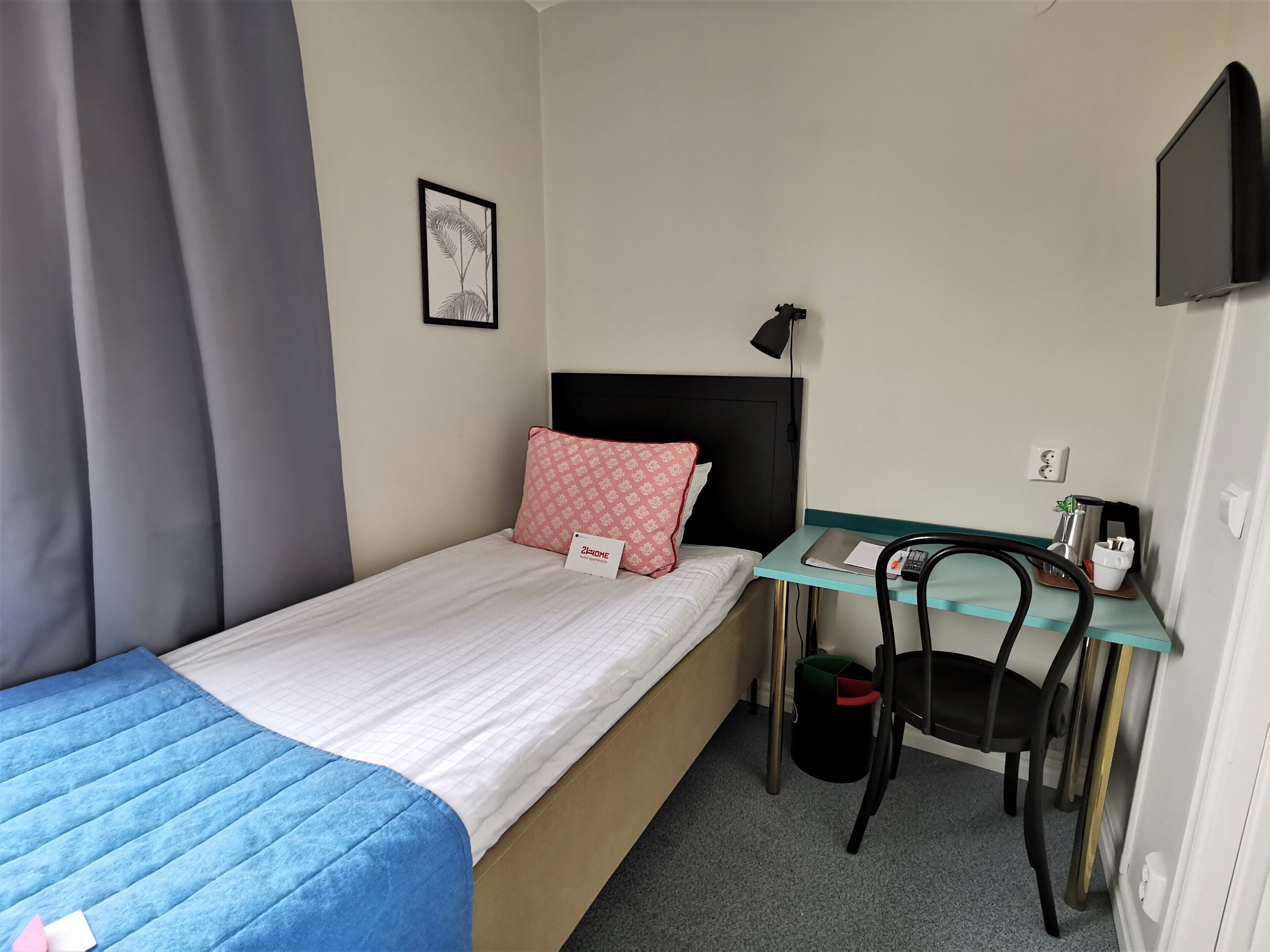 A small hotel room with a single bed, a desk with a chair, a TV mounted on the wall, and a small trash bin.