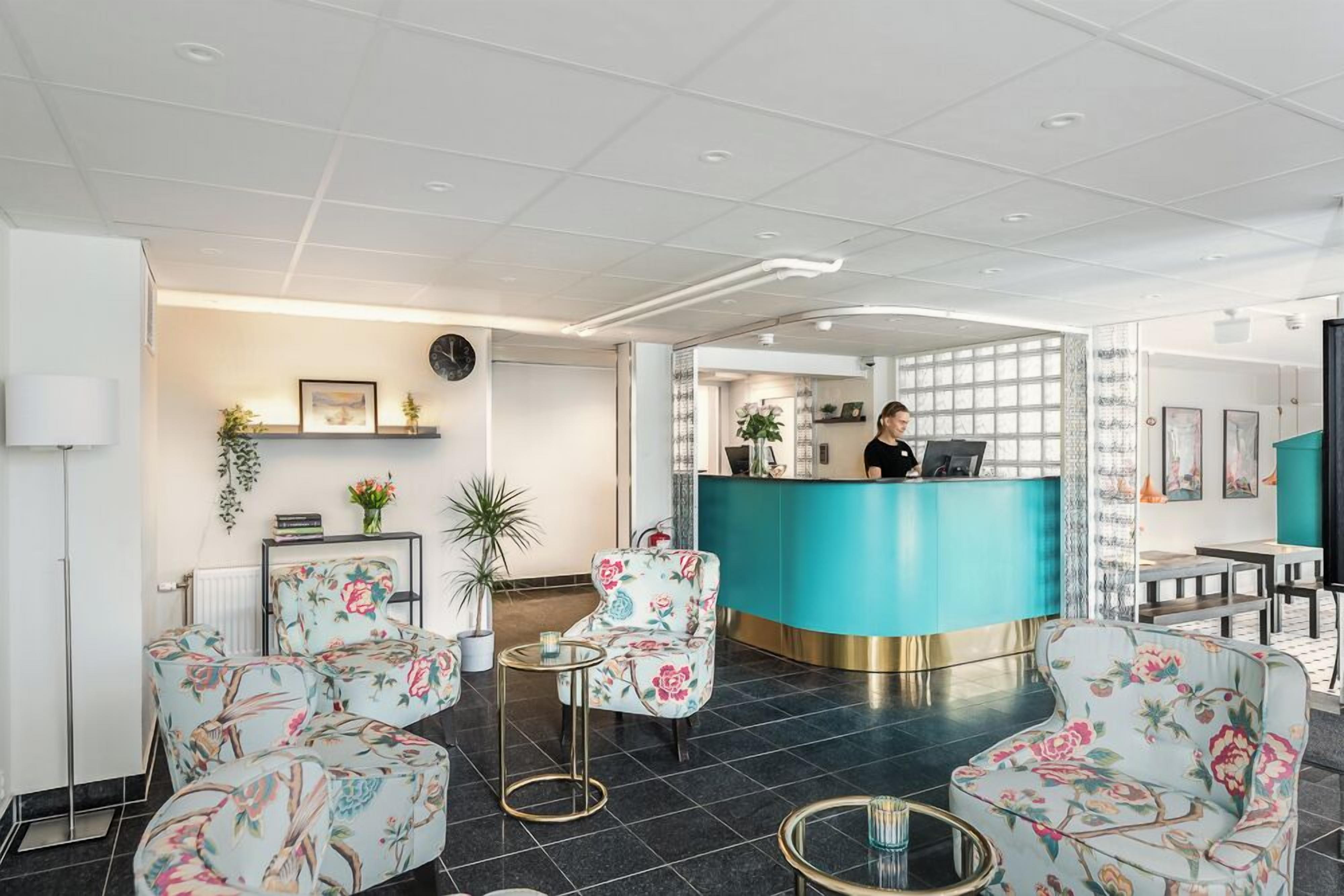 A hotel lobby with a reception desk, floral armchairs, a coffee table, and a potted plant.
