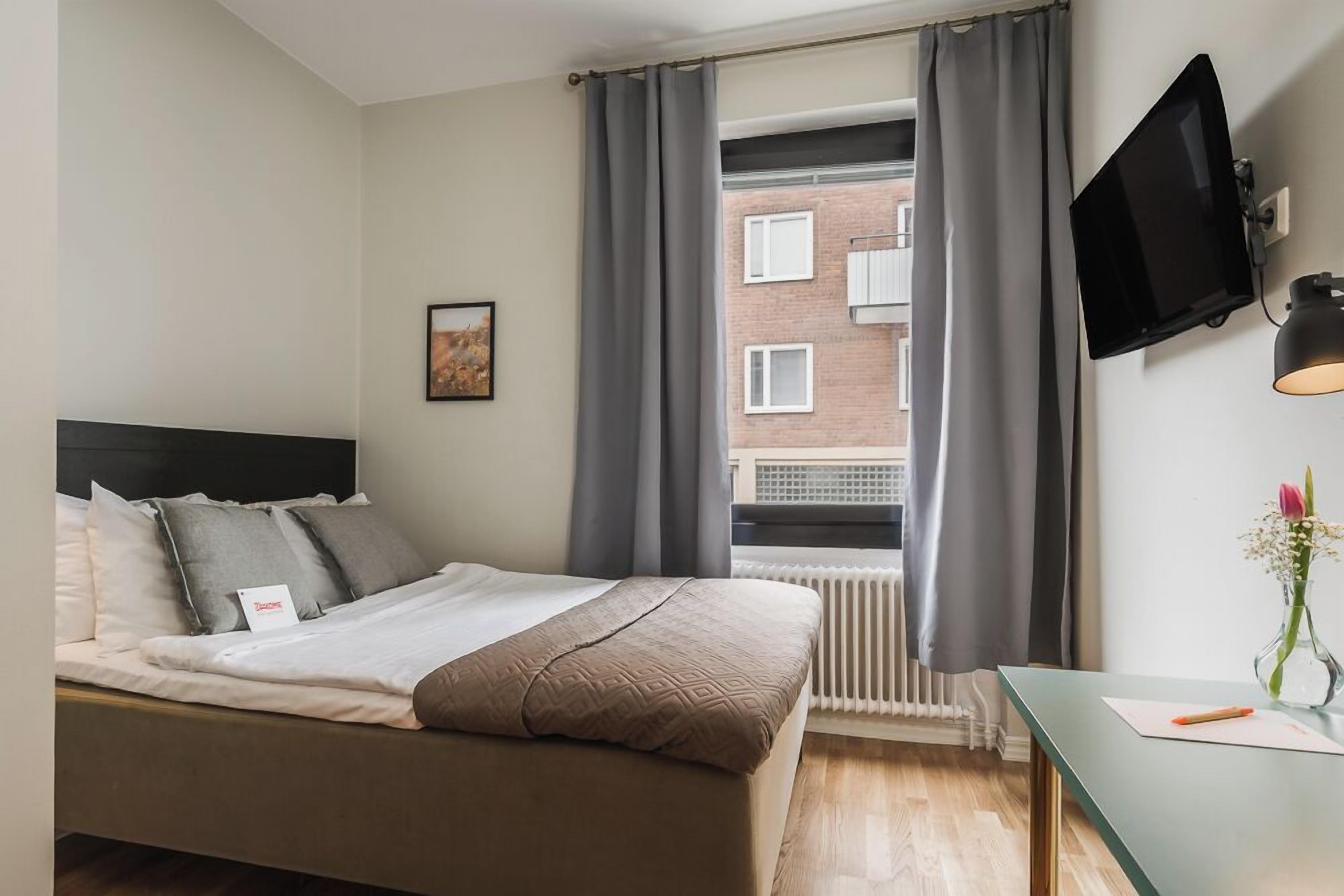 A bedroom with a bed, a window with curtains, a radiator, a TV mounted on the wall, and a small table with a vase.