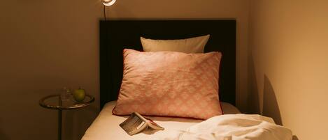 A neatly made bed with a single pillow and a book, a bedside table with a glass of water and an apple, and a framed picture on the wall.
