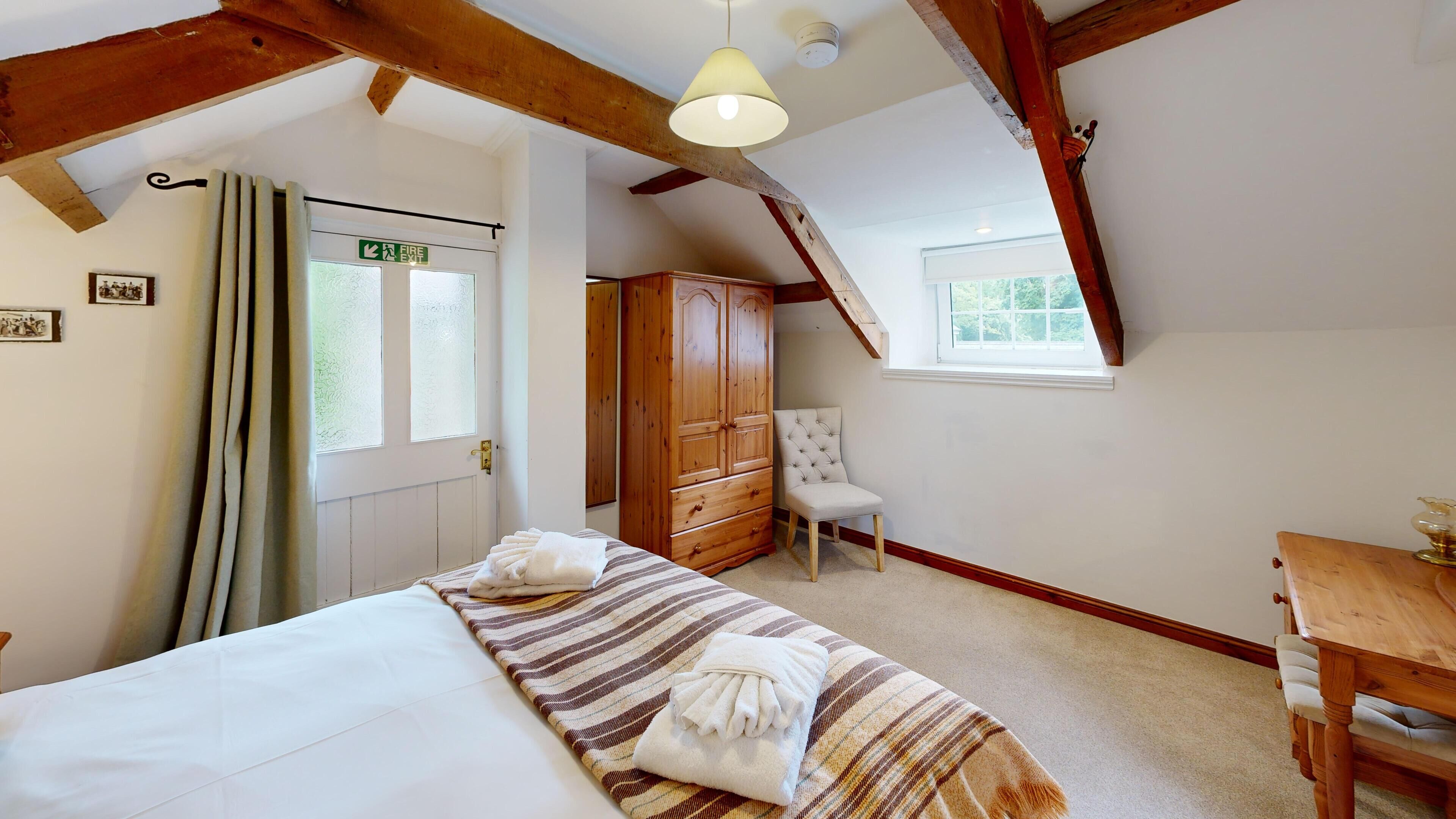 A bedroom with a bed, a wooden wardrobe, a chair, a small table, and a window.