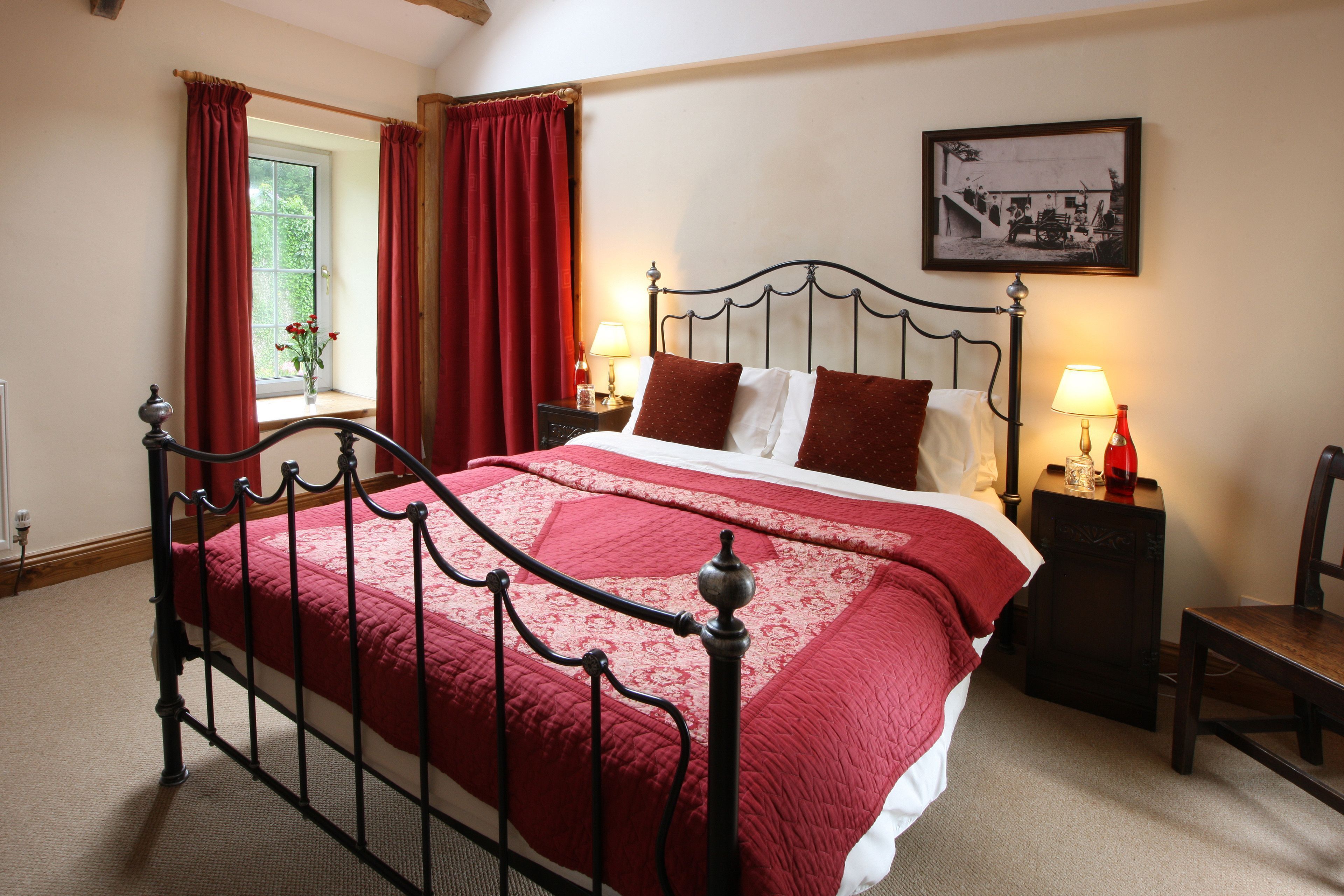 A neatly made bed with a red bedspread, a metal headboard, two bedside tables with lamps, and a window with red curtains.