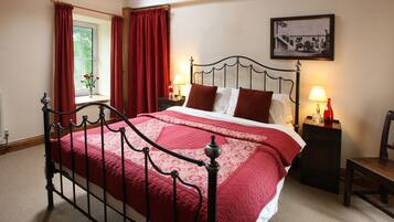A neatly made bed with a red bedspread, a metal headboard, two bedside tables with lamps, and a window with red curtains.