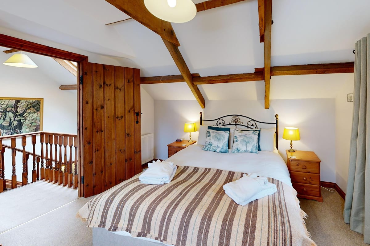 Een slaapkamer met een groot bed, houten balken, twee nachtkastjes met lampen, een houten kledingkast en uitzicht op een stadsbeeld door een raam.