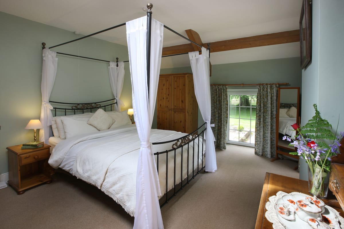 Een bed met hemelbed en witte beddengoed, een houten nachtkastje met een lamp, en een houten garderobe in een kamer met groene wanden en een houten plafondbalk.
