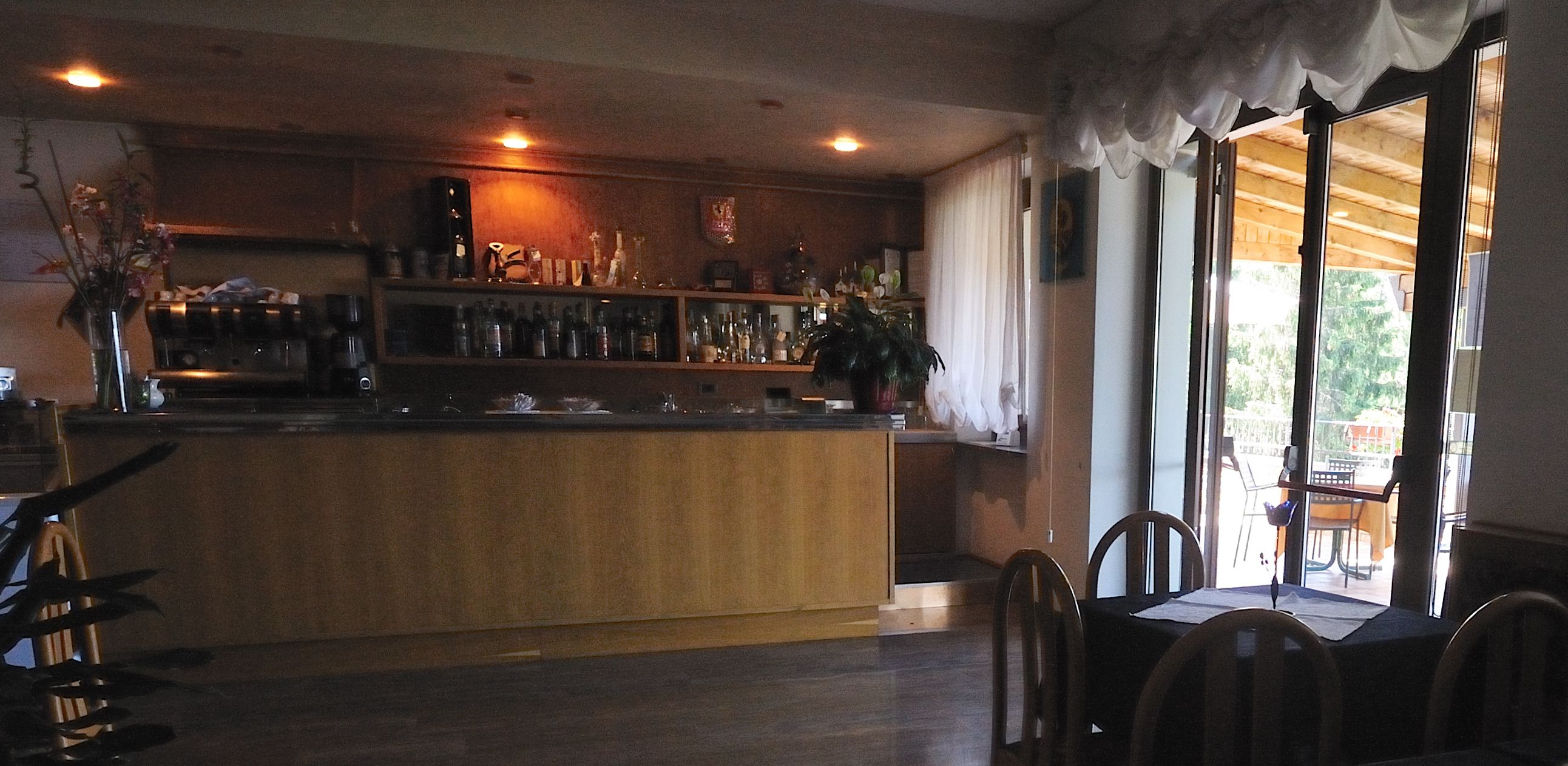 A bar area with a wooden counter, shelves with bottles, and a view of the outdoors through large windows.