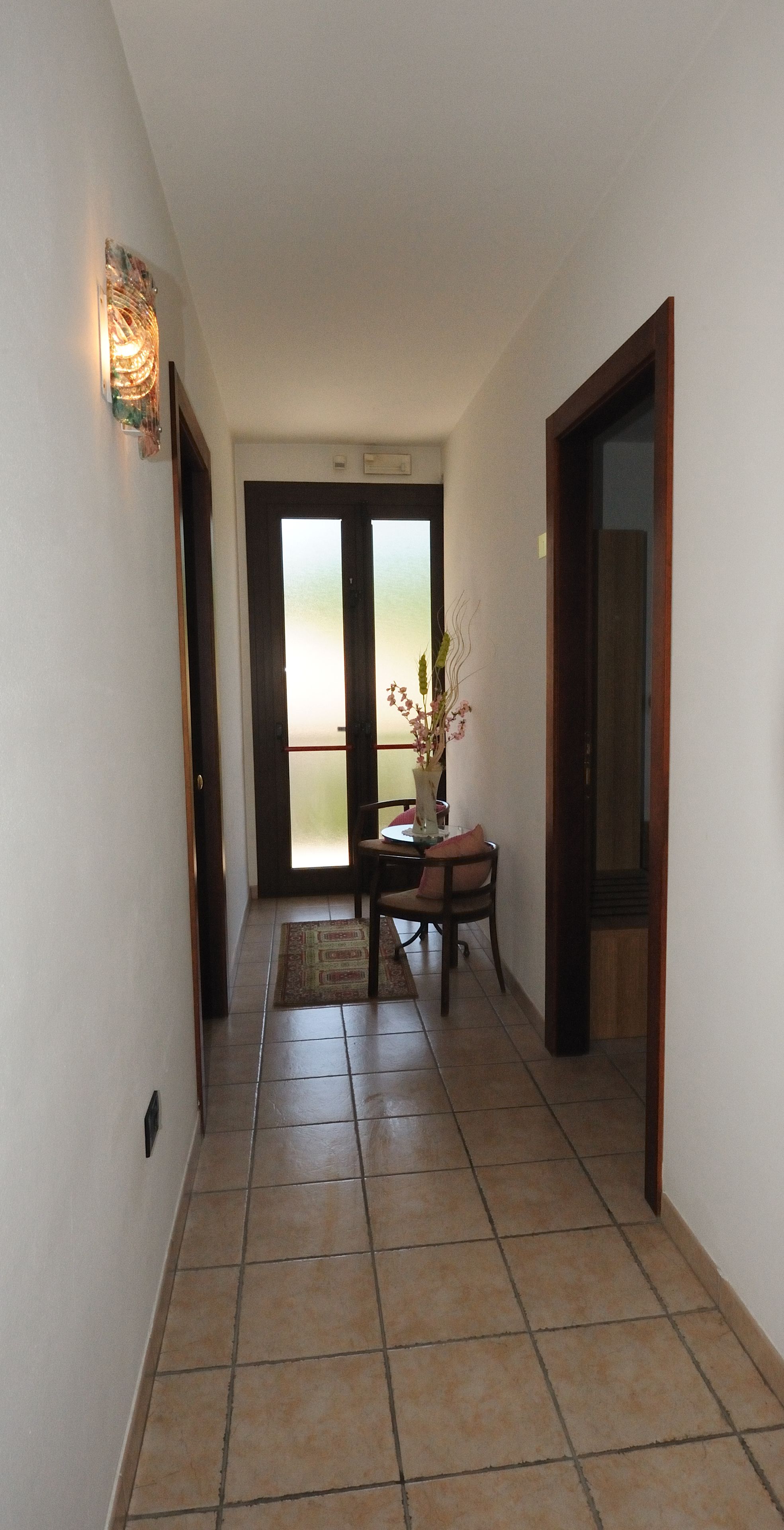 A hallway with tiled flooring, a dining area with a table and chairs, and a view through a glass door.
