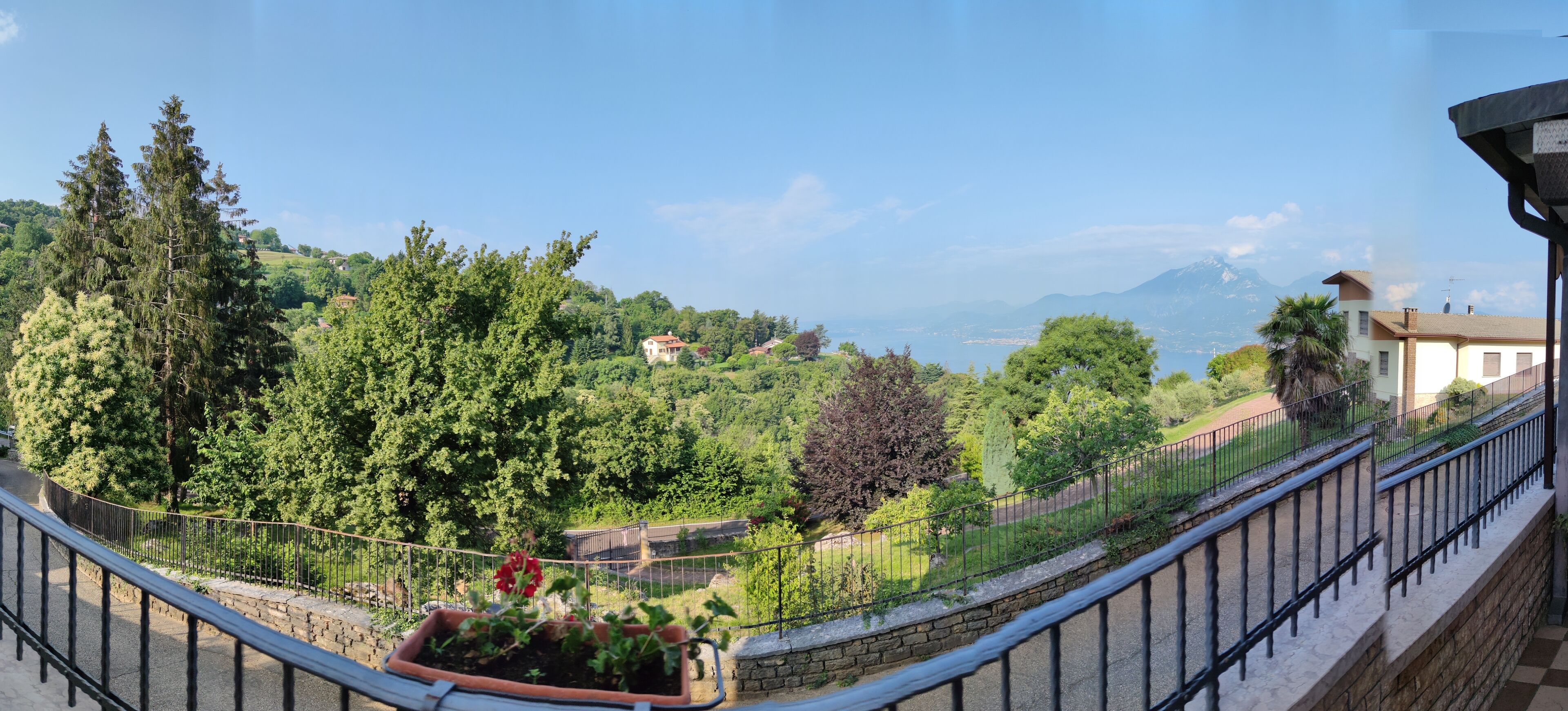 A balcony with a stone wall, a metal railing, and a view of a mountainous landscape with trees and buildings.