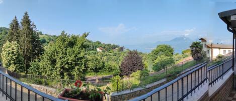 A balcony with a stone wall, a metal railing, and a view of a mountainous landscape with trees and buildings.
