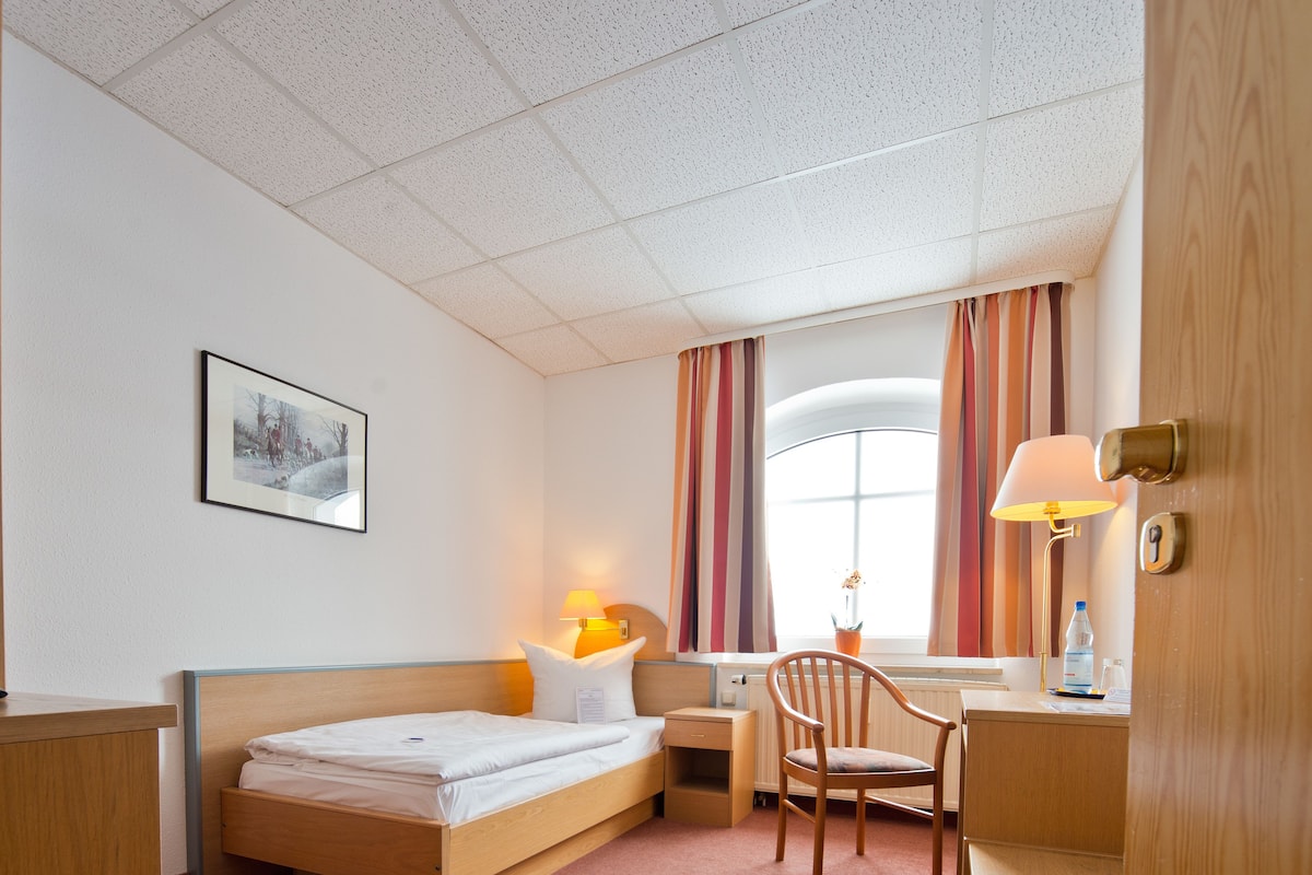 A hotel room with a bed, a desk, a chair, a window with curtains, a lamp, and a picture on the wall.