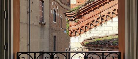 A balcony with wrought iron railings, a potted plant, and a view of a narrow street with historic buildings and a bell tower.