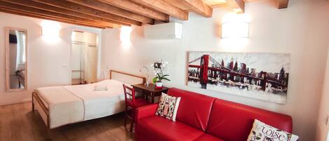 A room with a bed, a red sofa, a dining table, and a painting of a bridge.