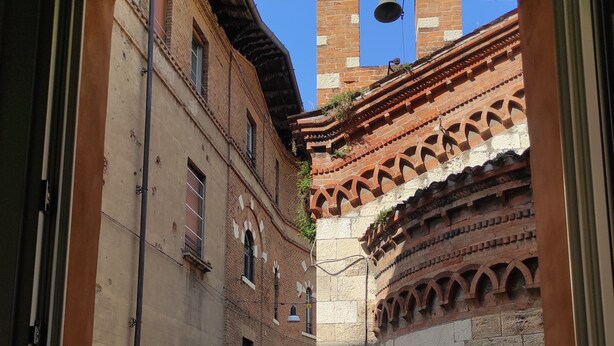 A narrow alleyway with buildings on either side, a balcony with a wrought iron railing, and a potted plant with flowers. - B&B Ai Musei (Brescia)