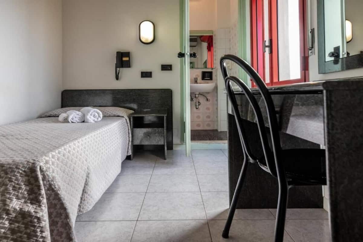 Habitación de hotel con cama, escritorio, silla, baño a la vista a través de una puerta abierta y un espejo en la pared.