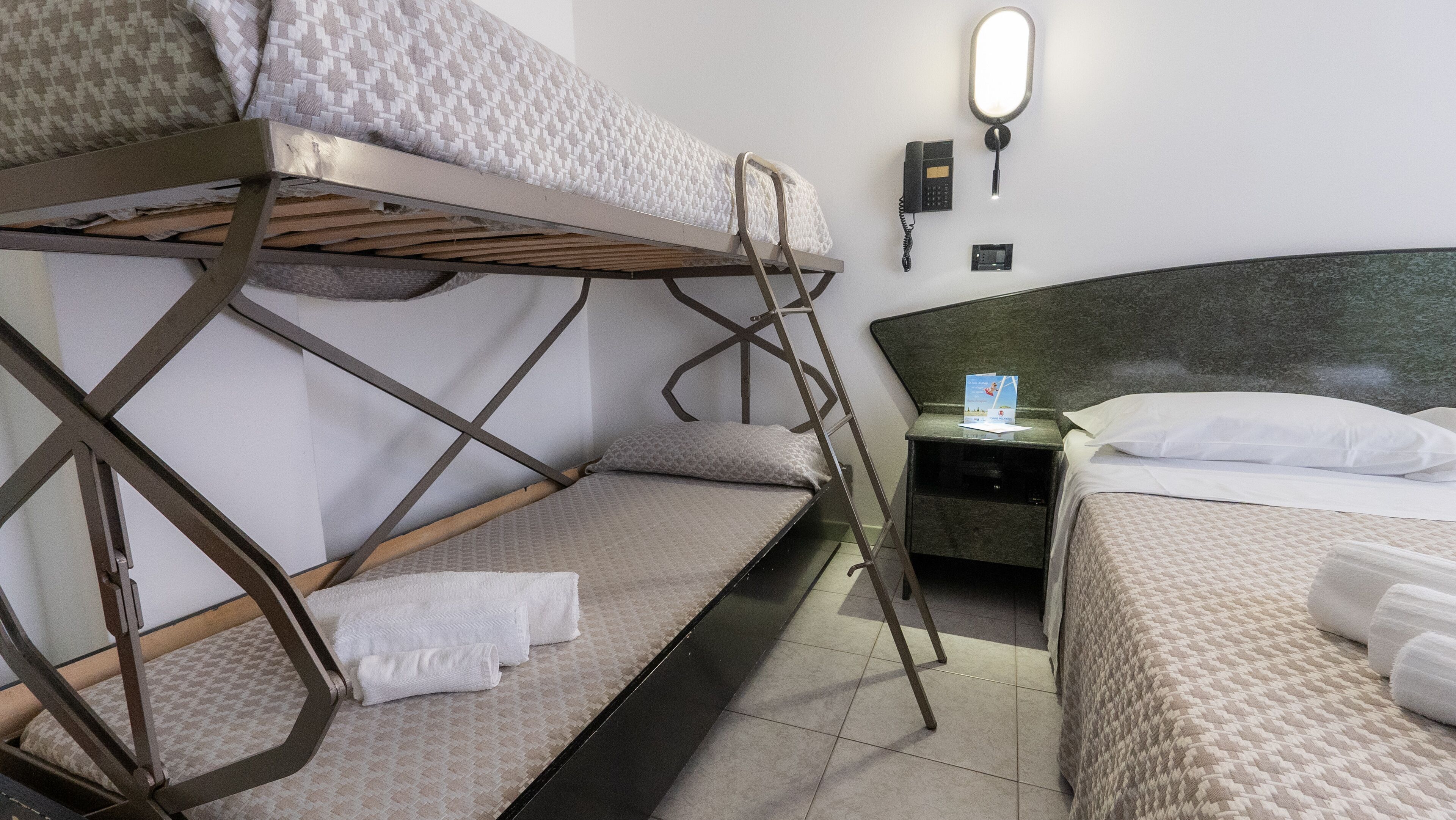A hotel room with bunk beds, a bedside table with a lamp, and a wall-mounted phone.
