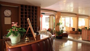A lobby with a wooden reception desk, a clock, and a seating area with sofas and potted plants. - Hotel Nobel (Rimini)