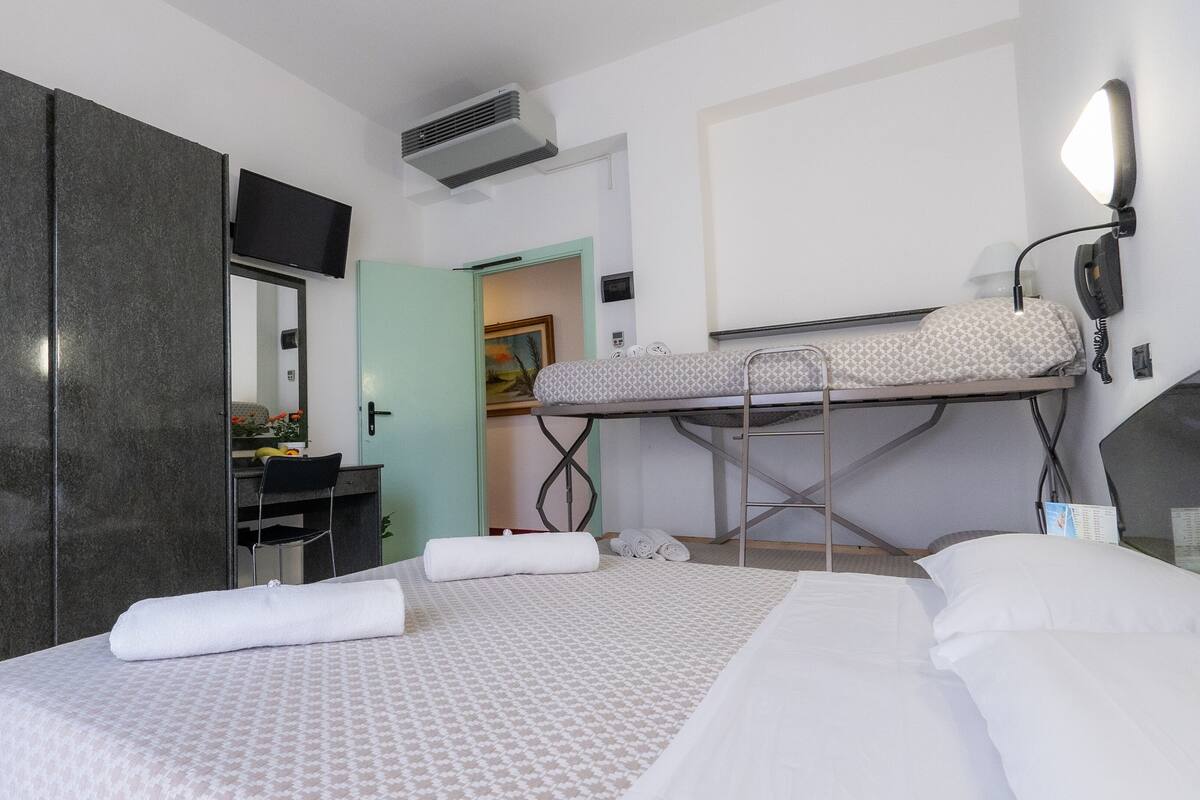 Una habitación de hotel con una cama, un armario, un escritorio con silla, una lámpara colgante y una litera con escalera.