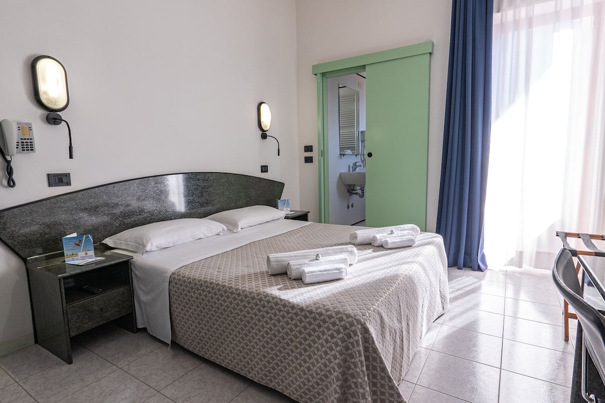 Habitación de hotel con cama, mesita de noche y velador. Hay un teléfono en la pared y una puerta con marco verde.