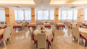 A spacious dining area with multiple round tables, each set with white tablecloths and red runners. The room has large windows with sheer curtains, allowing natural light to fill the space. - Hotel Nobel (Rimini)