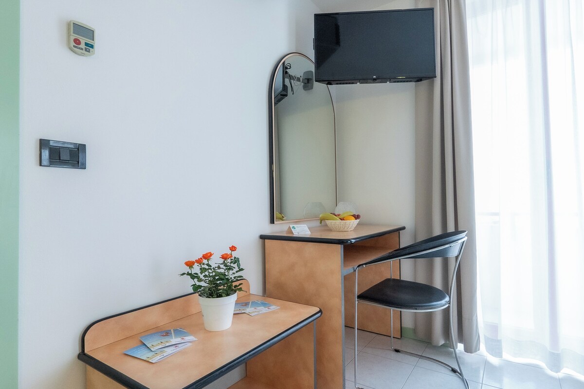Habitación de hotel con escritorio de madera, una silla, una pequeña planta en maceta, una cesta de frutas, un televisor de pantalla plana, un espejo y una ventana con cortinas.
