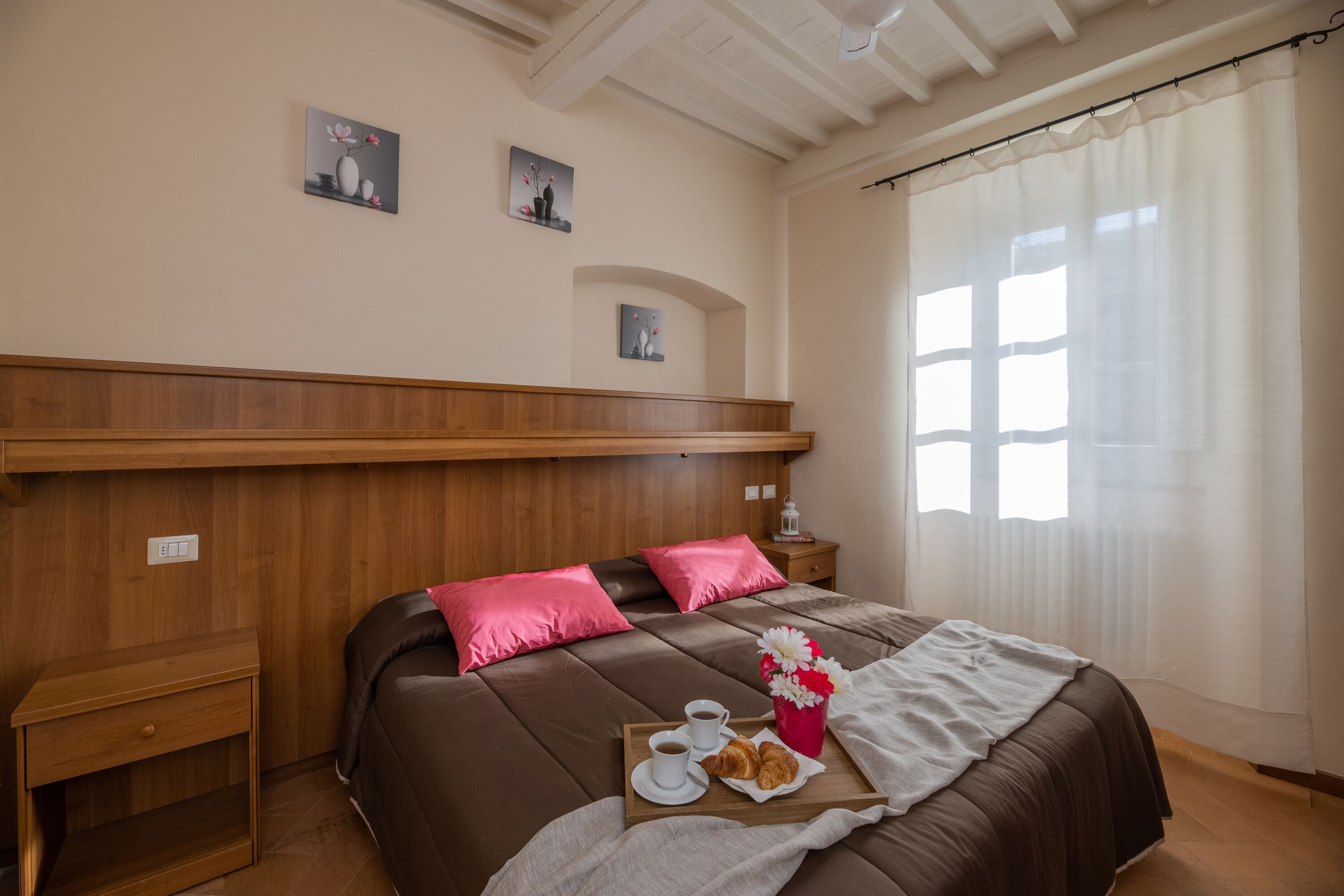 Quarto bem arrumado com cabeceira de madeira, cama com roupa de cama rosa e marrom, mesa de cabeceira com abajur e bandeja com itens de café da manhã.