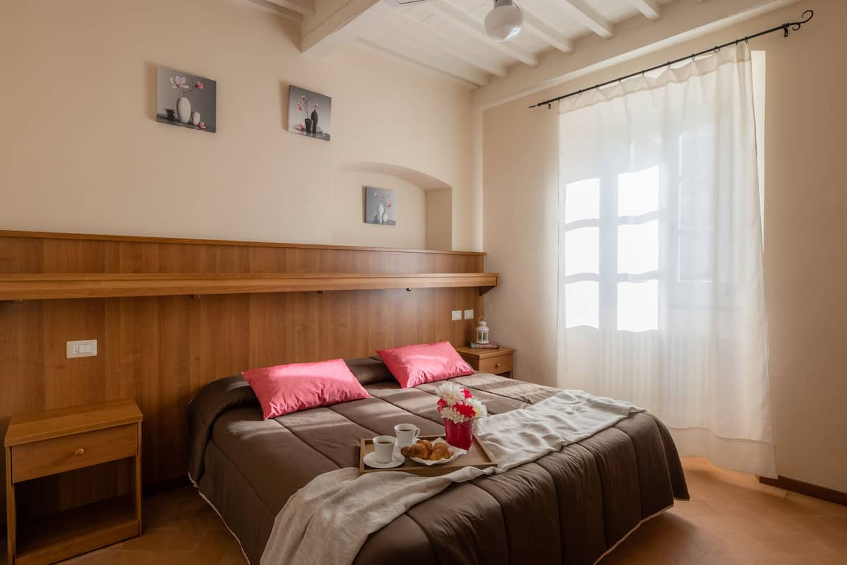 A bedroom with a wooden headboard, a bed with pink pillows, a bedside table with a lamp, and a window with sheer curtains.