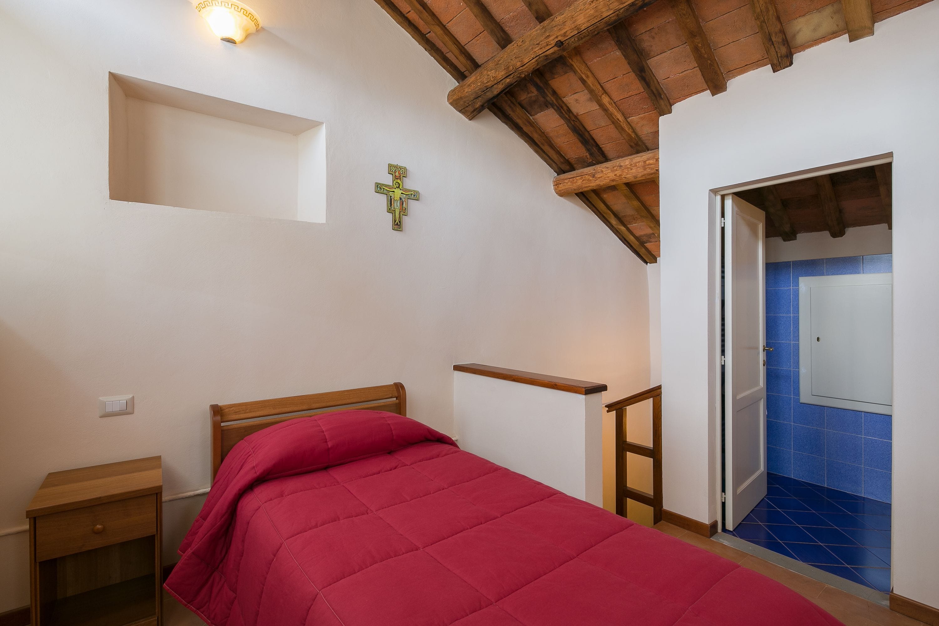 A bedroom with a wooden bed, a nightstand, a cross on the wall, and a door leading to a bathroom with blue tiles.
