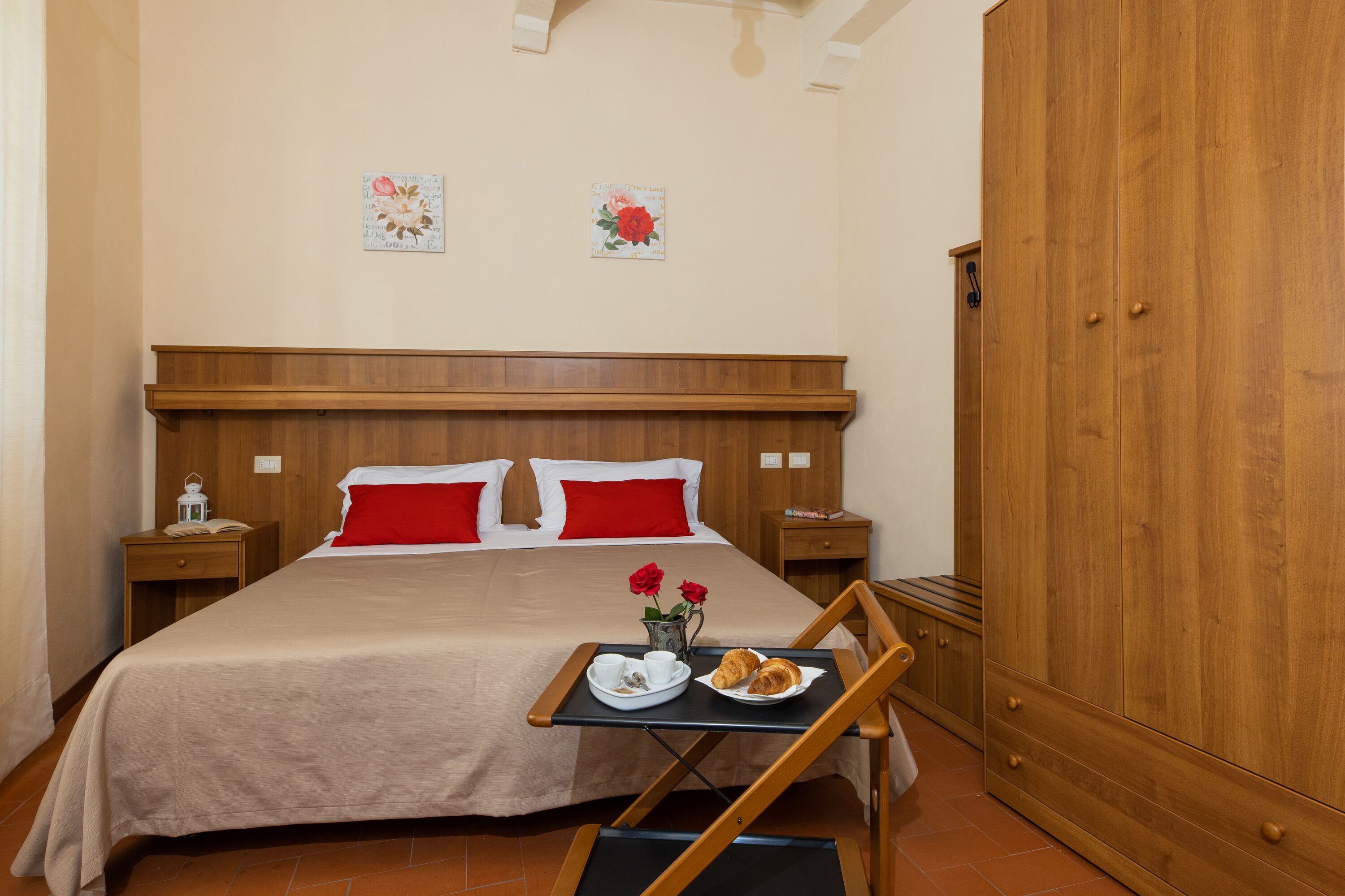 Ein ordentlich bezogenes Bett mit roten Kissen, ein Nachttisch mit Lampe, ein Holzschrank und ein Tablett mit Croissants und Blumen auf dem Boden.