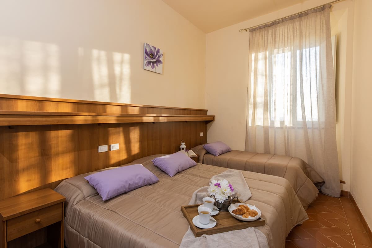 A hotel room with two beds, a wooden headboard, a nightstand, a tray with breakfast items, and a window with sheer curtains.