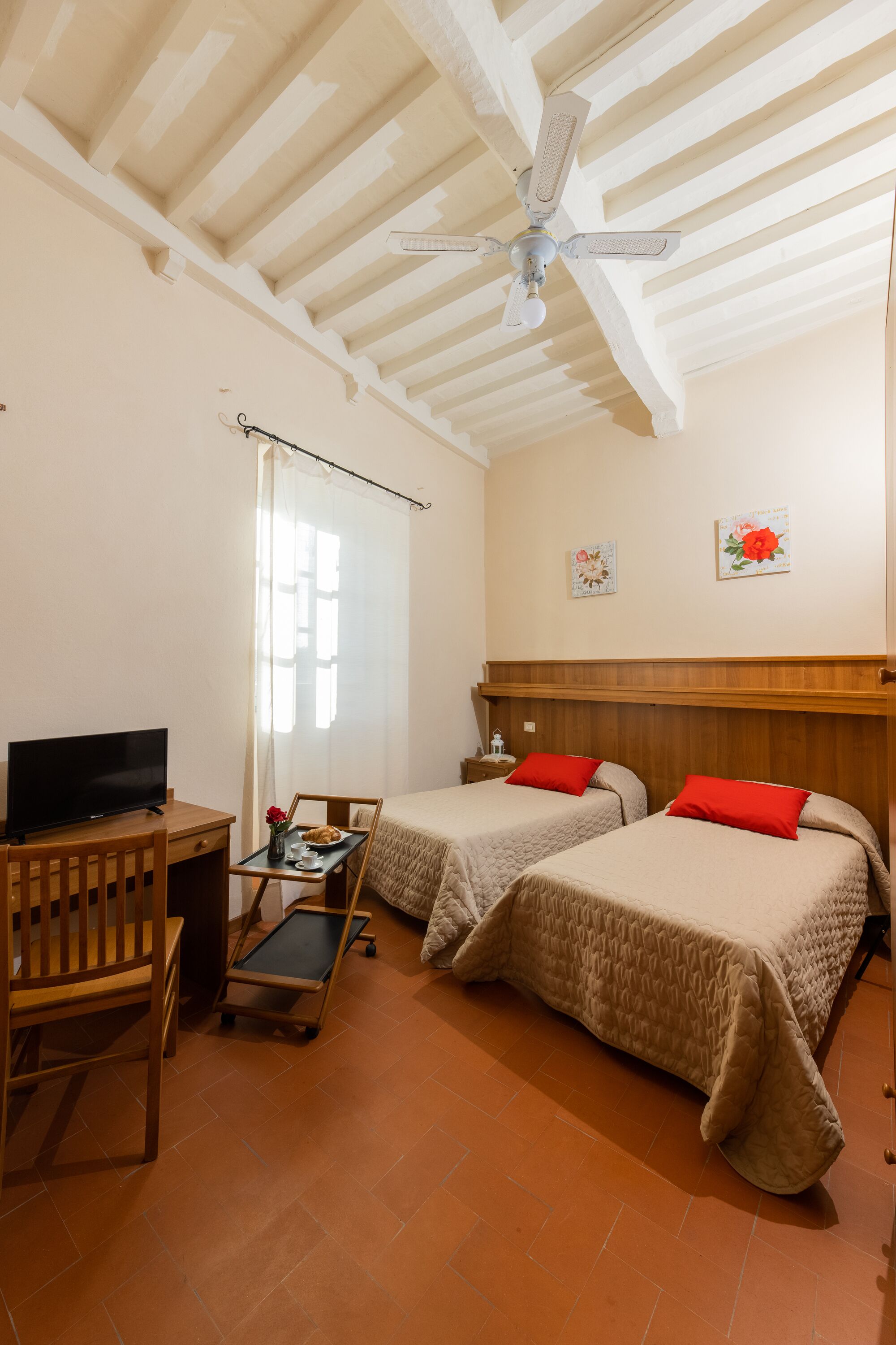 A room with two beds, a desk with a TV, a ceiling fan, and a window with curtains.