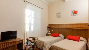 A room with two beds, a desk with a TV, a ceiling fan, and a window with curtains.