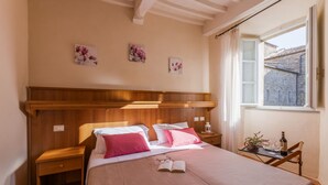 Un dormitorio con cabecera de madera, dos camas con ropa de cama rosa y blanca, una mesita de noche con lámpara, una pequeña mesa con una botella y vasos, y una ventana con vistas a edificios.
