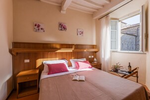 Egy hálószoba, melyben egy fafejű ágy található, két ágy rózsaszín és fehér ágyneművel, egy éjjeliszekrény lámpával, egy kis asztal egy üveggel és poharakkal, valamint egy ablak, melyből épületekre nyílik a kilátás.