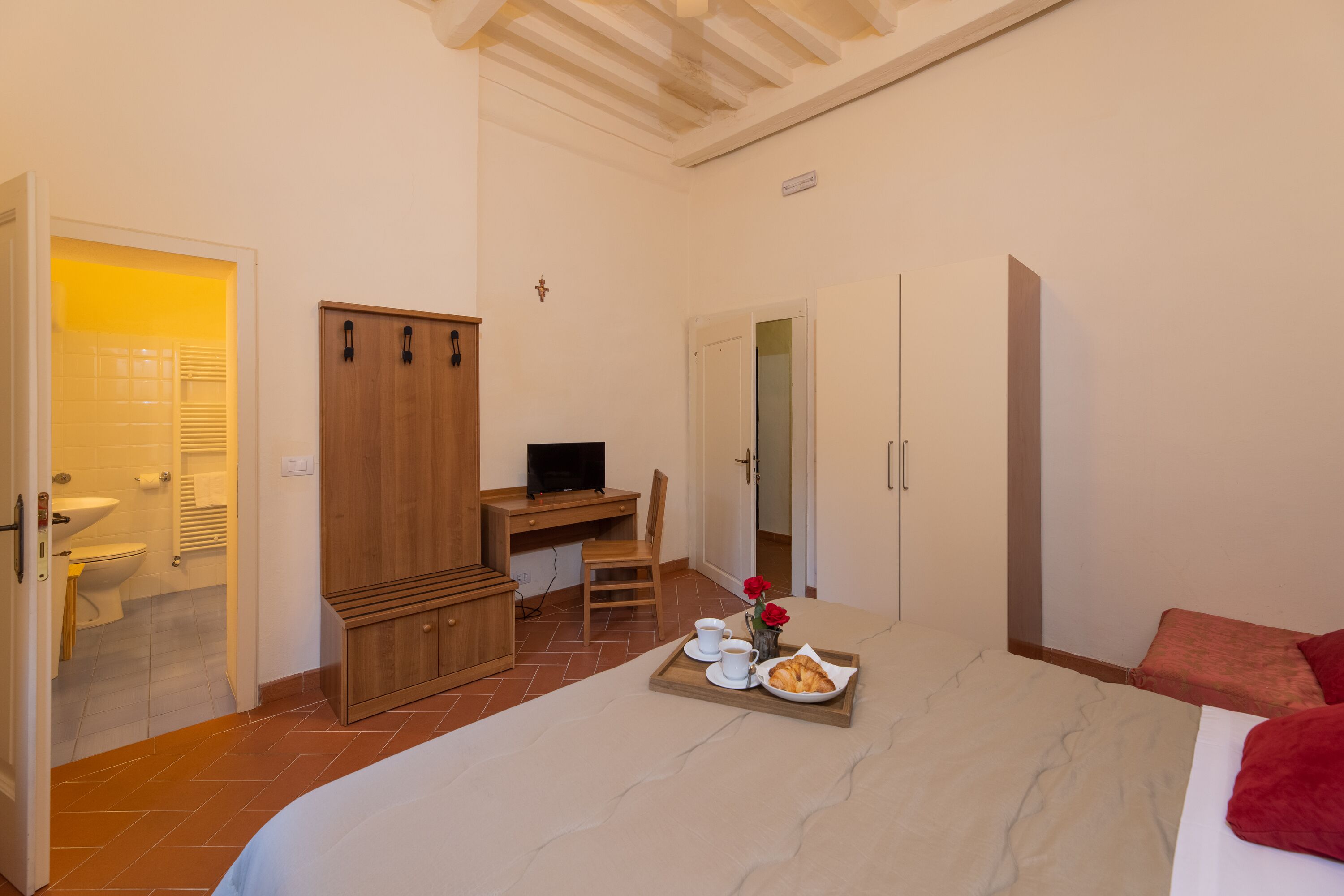 Ein Schlafzimmer mit einem Bett, einem Schreibtisch mit Computer, einem Kleiderschrank und einem Tablett mit Croissant und Kaffee.