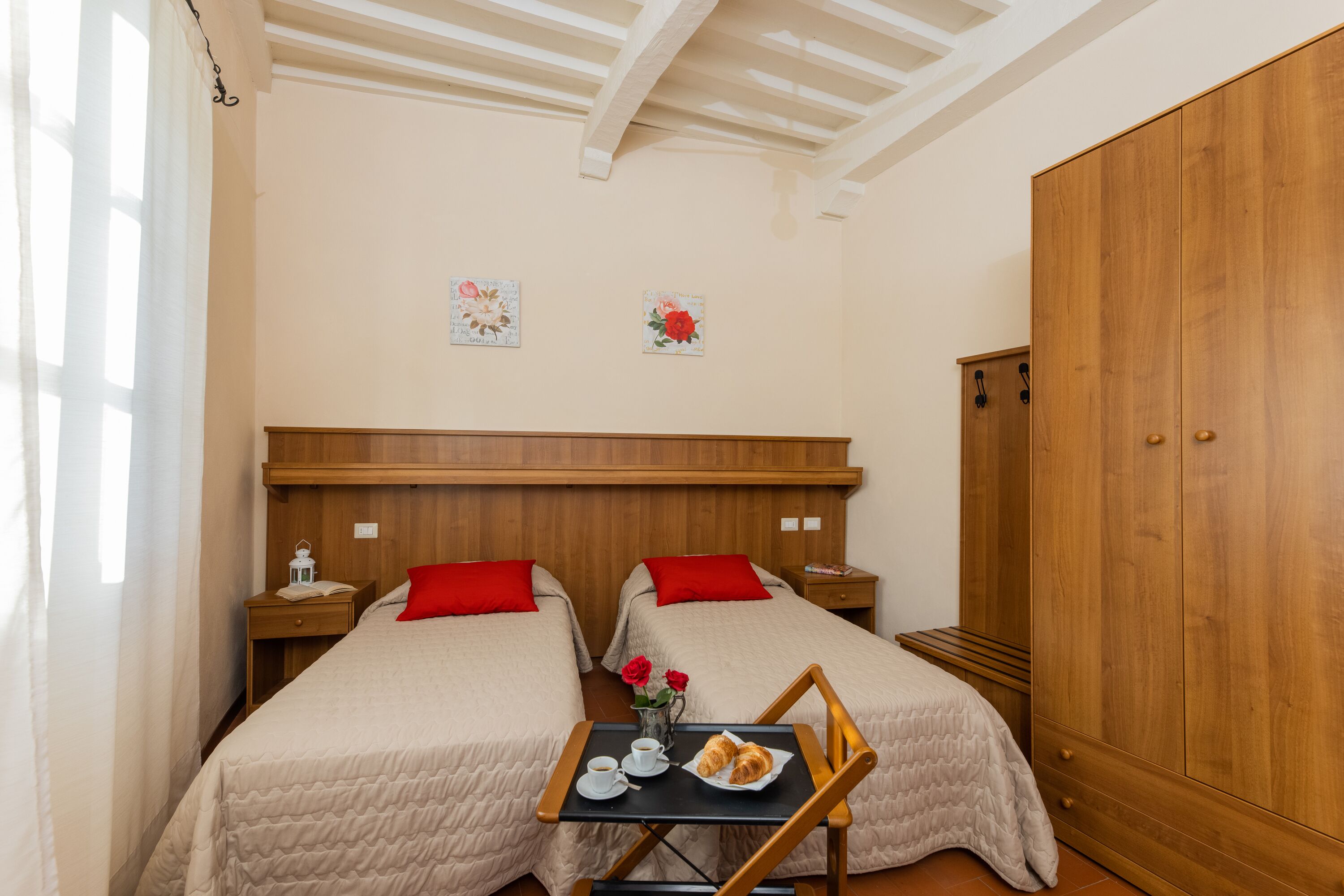 Una habitación con dos camas, un armario de madera, una mesita con una bandeja de cruasanes y una taza, y una mesita de noche con una lámpara.