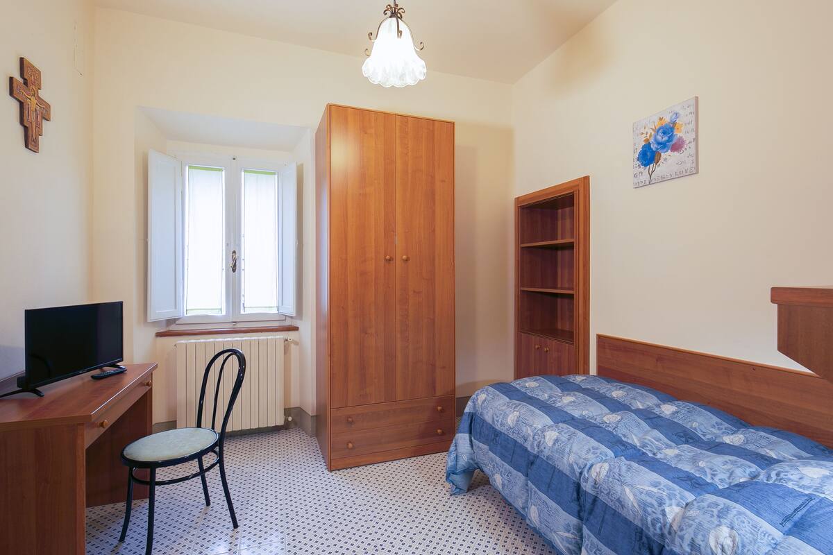 A bedroom with a wooden wardrobe, a bed with a blue patterned duvet, a desk with a television, and a chair.