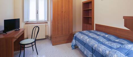 A bedroom with a wooden wardrobe, a bed with a blue patterned duvet, a desk with a television, and a chair.