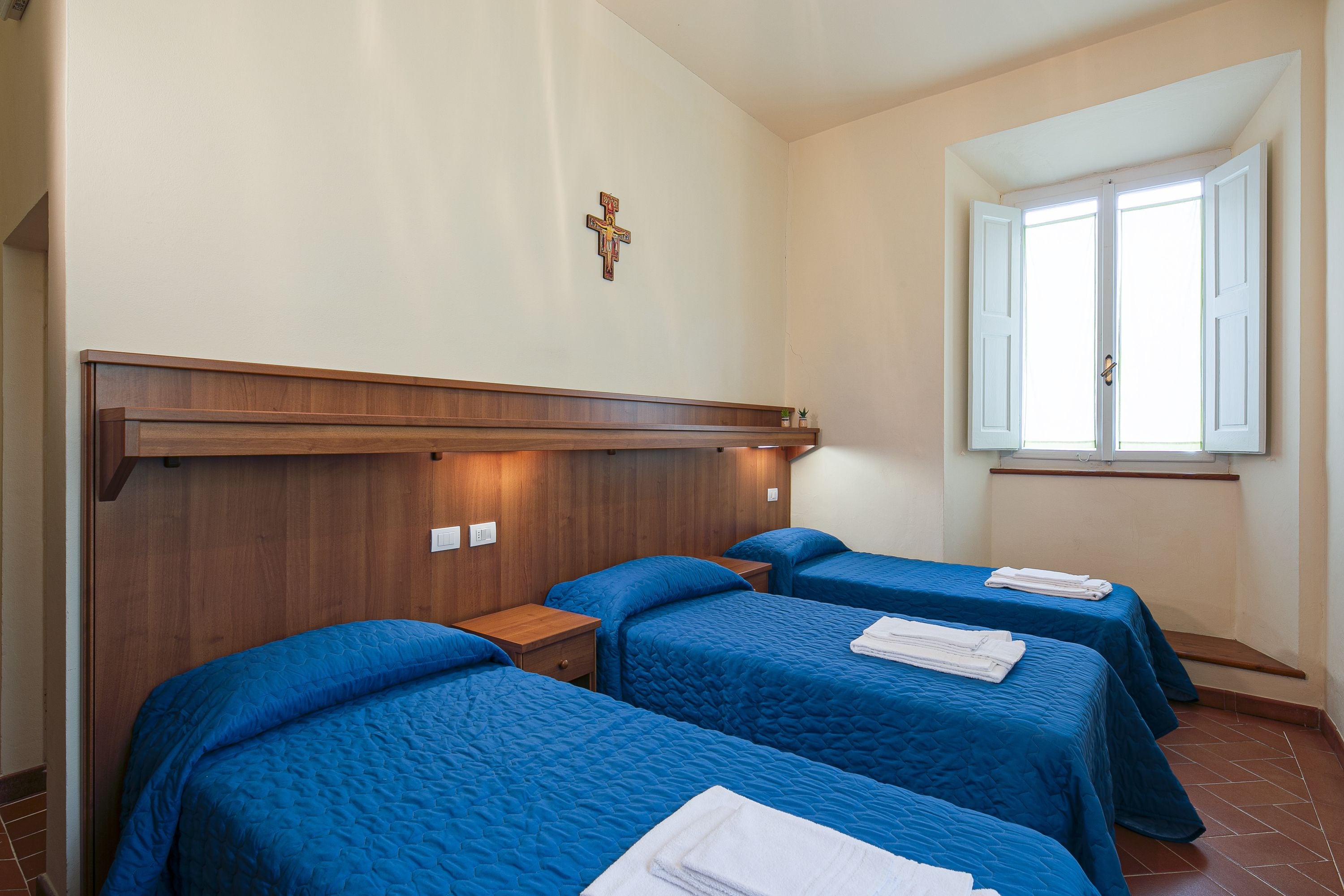 Habitación de hotel con tres camas, cabeceras de madera, una cruz en la pared y una ventana con contraventanas blancas.