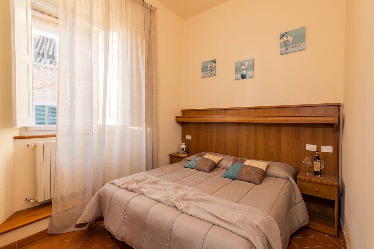 A bedroom with a wooden headboard, a bed with a beige and brown duvet, two bedside tables with lamps, and a window with a sheer curtain.