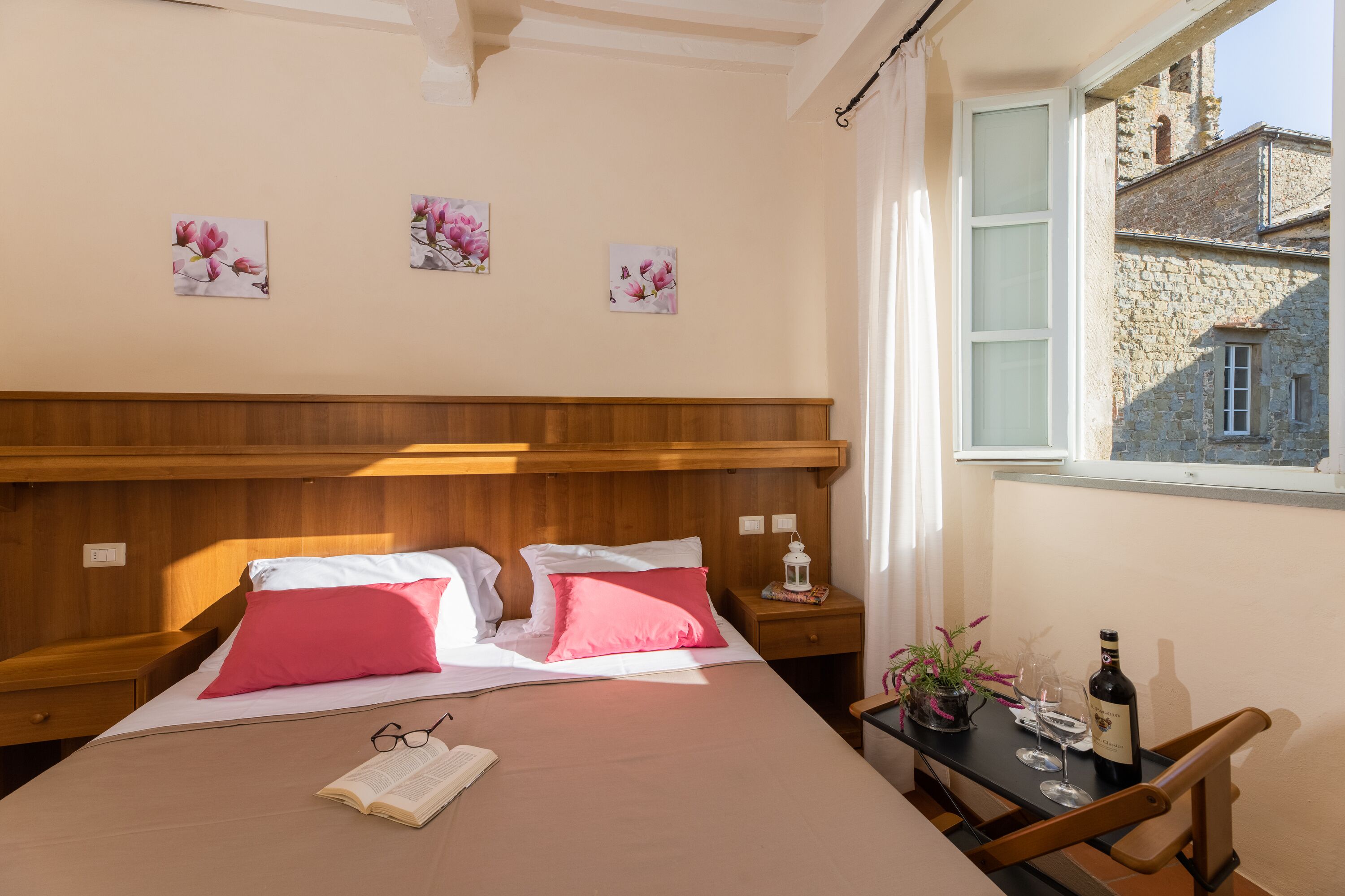 A bedroom with wooden headboard, a bed with pink and white pillows, a bedside table with a wine bottle and glasses, and a window with a view of stone buildings.