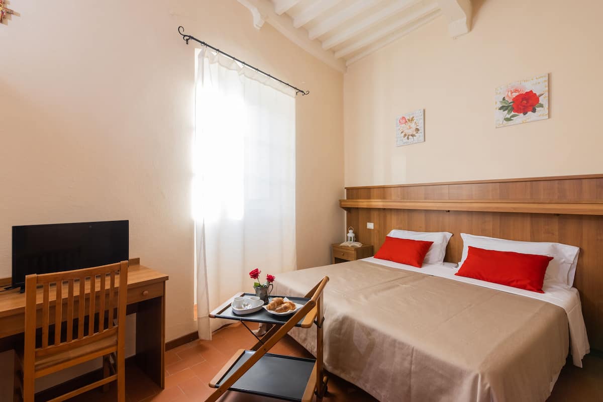 A hotel room with a wooden desk, a bed with red pillows, a TV on the desk, and a window with curtains.