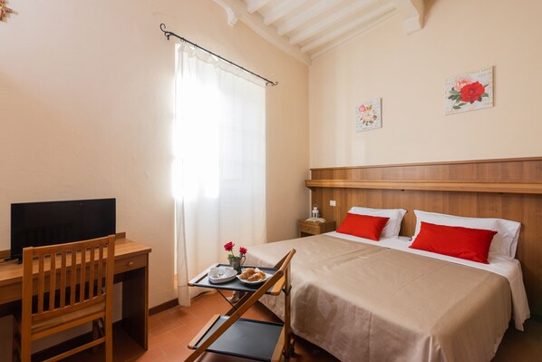 A hotel room with a wooden desk, a bed with red pillows, a TV on the desk, and a window with curtains. - B&B San Francesco (Cortona)