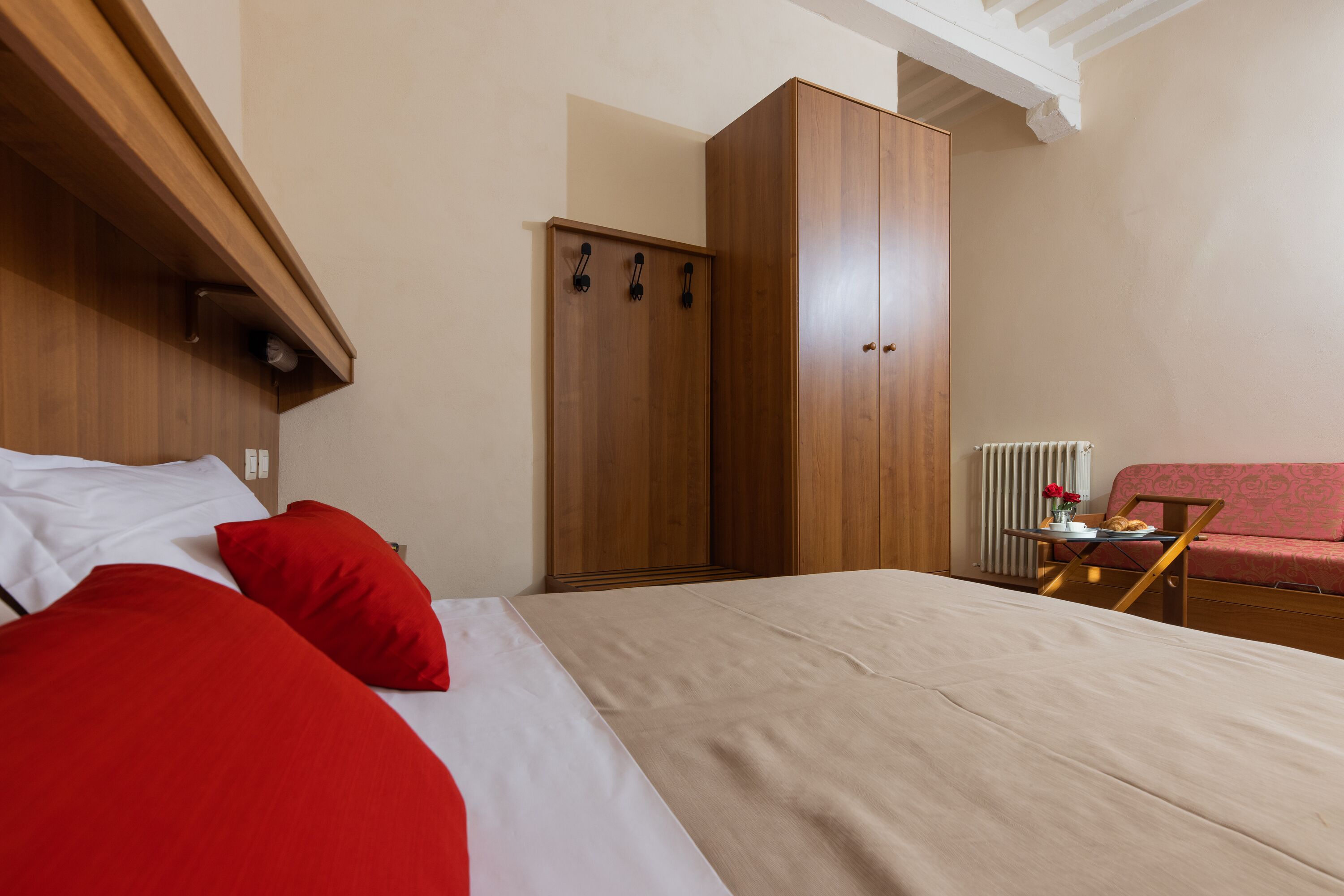 A bedroom with a bed, a wardrobe, a radiator, and a small table with a vase and flowers.