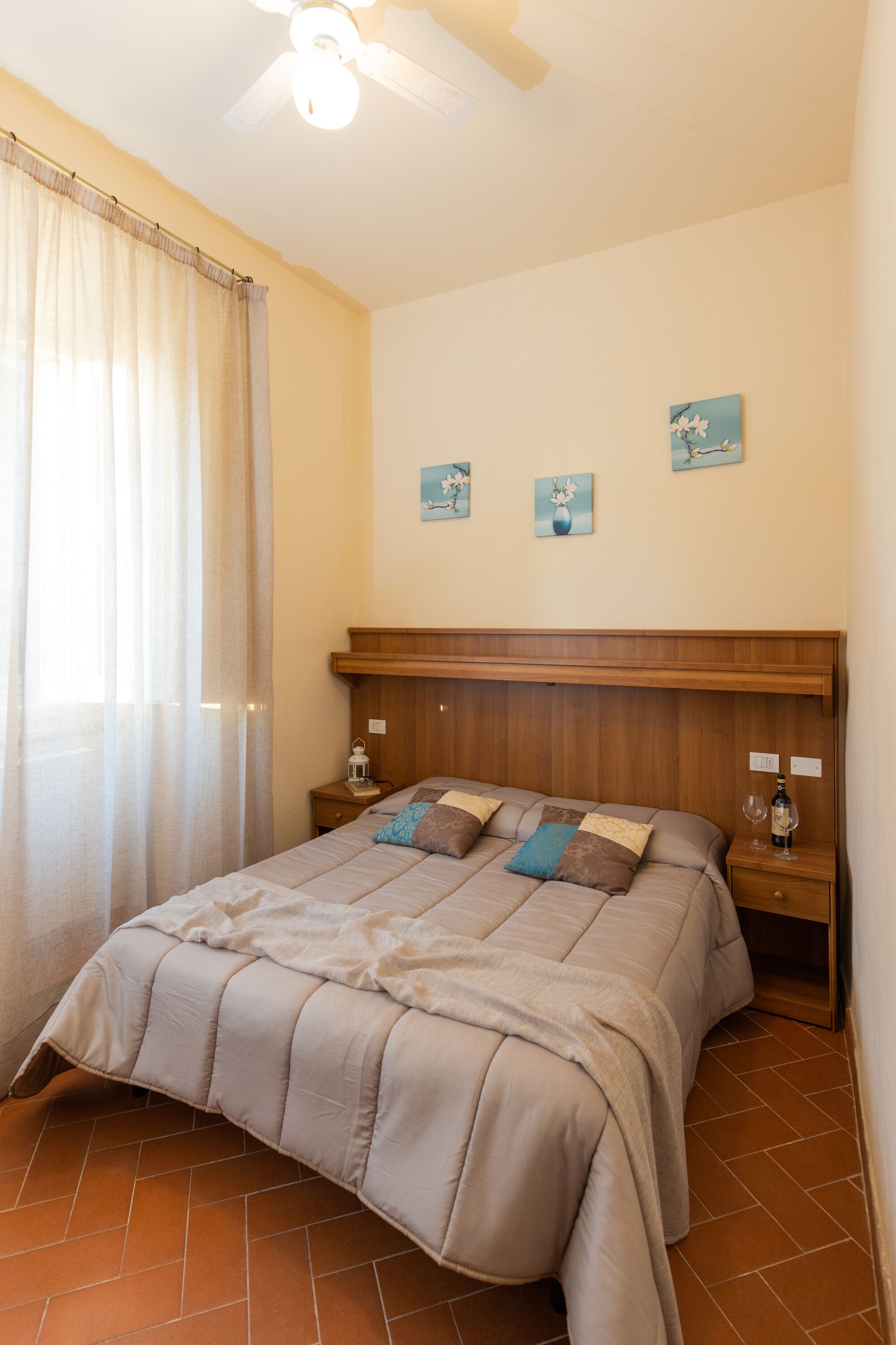 Quarto com cama, cabeceira de madeira, duas mesas de cabeceira, um criado-mudo, um ventilador de teto e janela com cortinas.