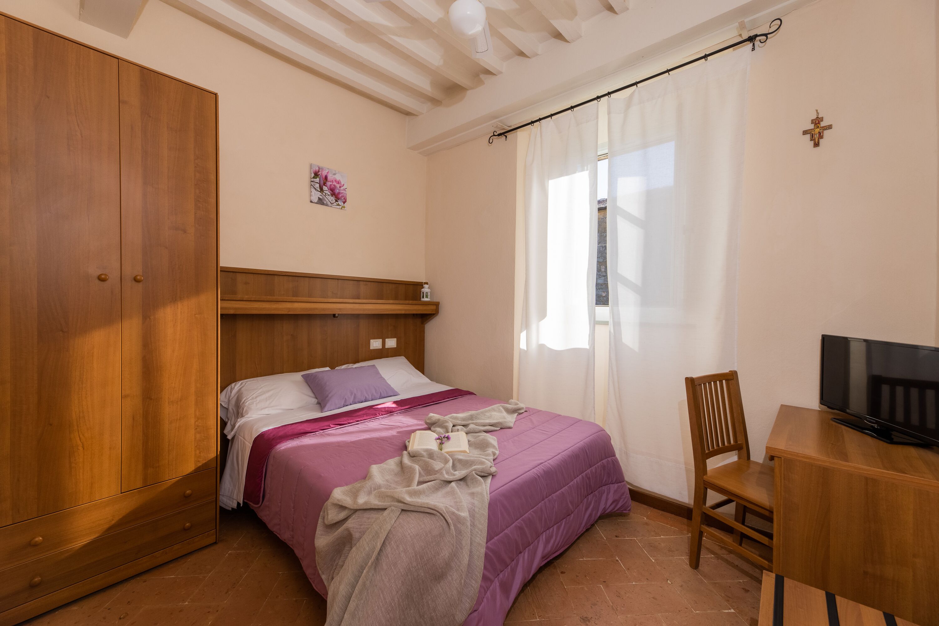 A bedroom with a bed, wooden wardrobe, a desk with a television, and a window with curtains.