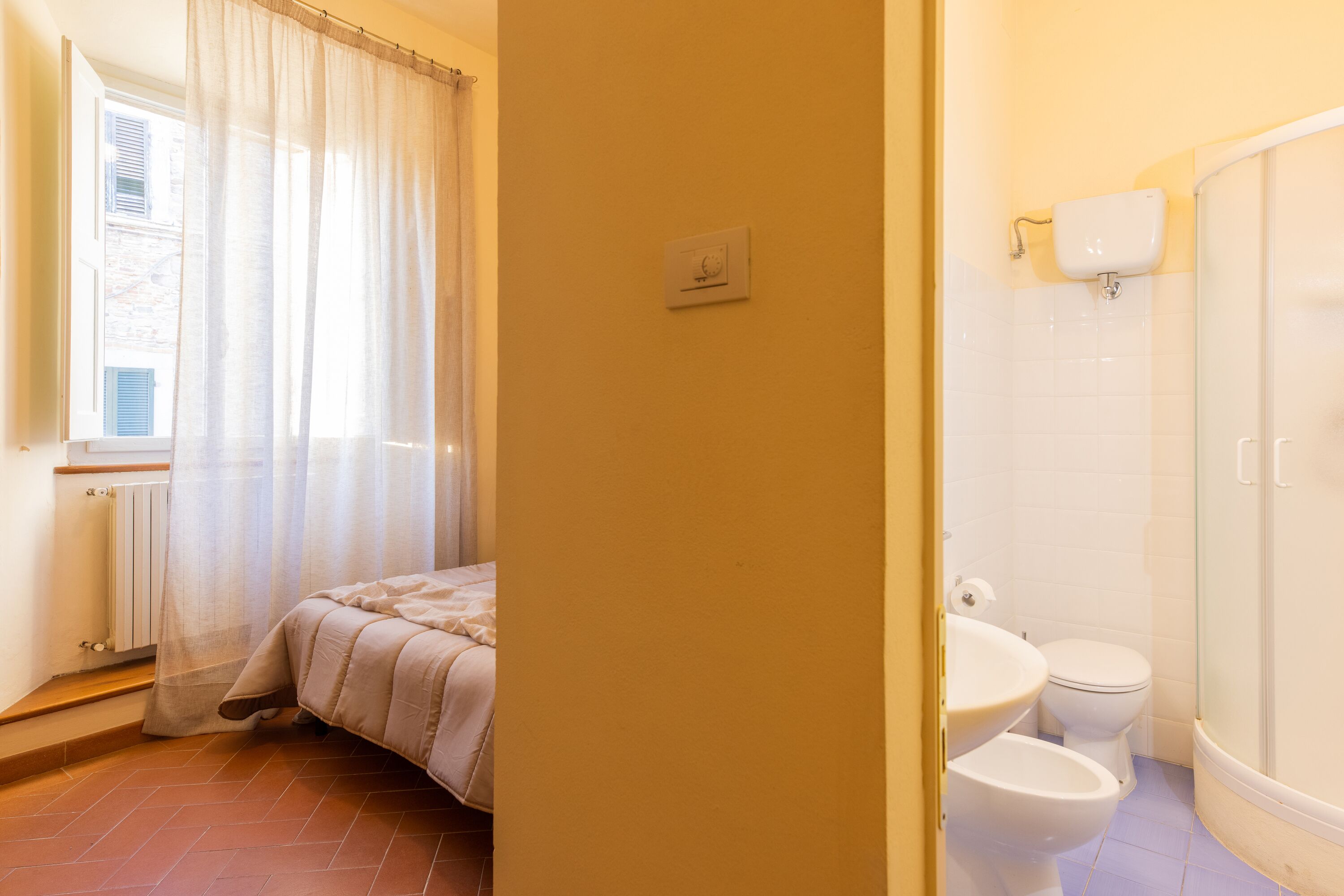 A bedroom with a bed, a bathroom with a toilet and a shower, and a window with curtains.