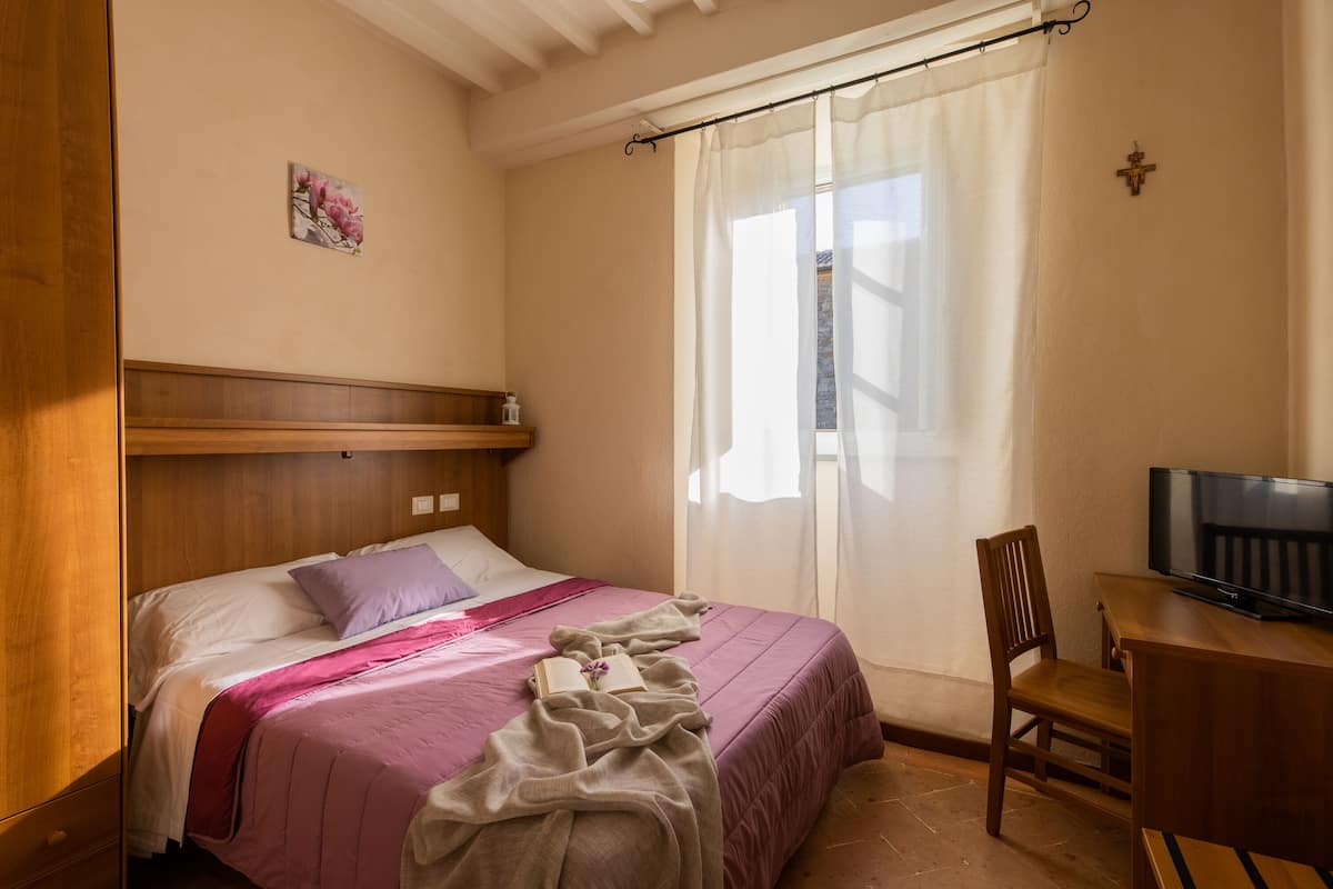 A bedroom with a bed, a wooden headboard, a television on a wooden table, and a window with curtains.