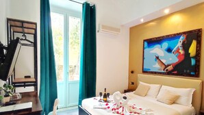 A hotel room with a bed, a desk, a chair, a painting on the wall, and a window with curtains. - Chroma Italy - Chroma Deka (Rome)
