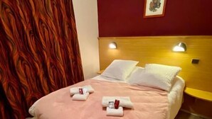 Una habitación de hotel con cama, dos almohadas, cabecero, mesita de noche, lámpara de pared y cortina.