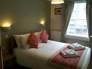 A neatly made bed with white and red pillows, a bedside table with a lamp, and a window with curtains. - The Crown Hotel (Corwen)